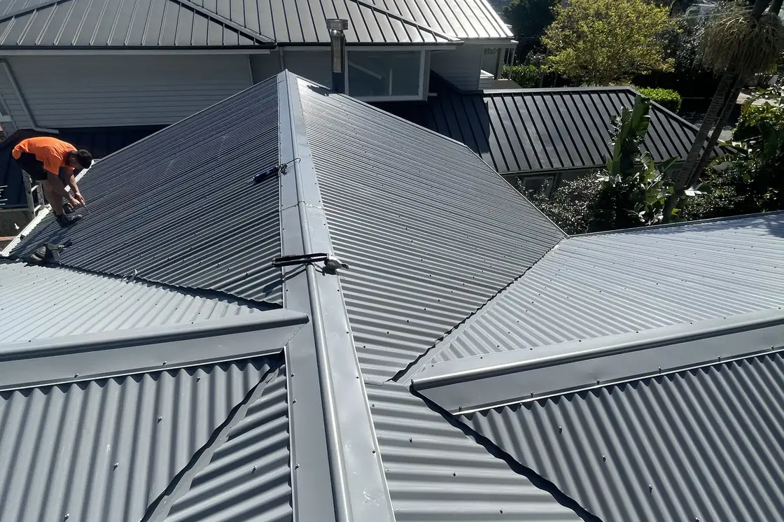 A complex roof of hips and valleys in new steel, a roofer working at the far ridge and trees behind