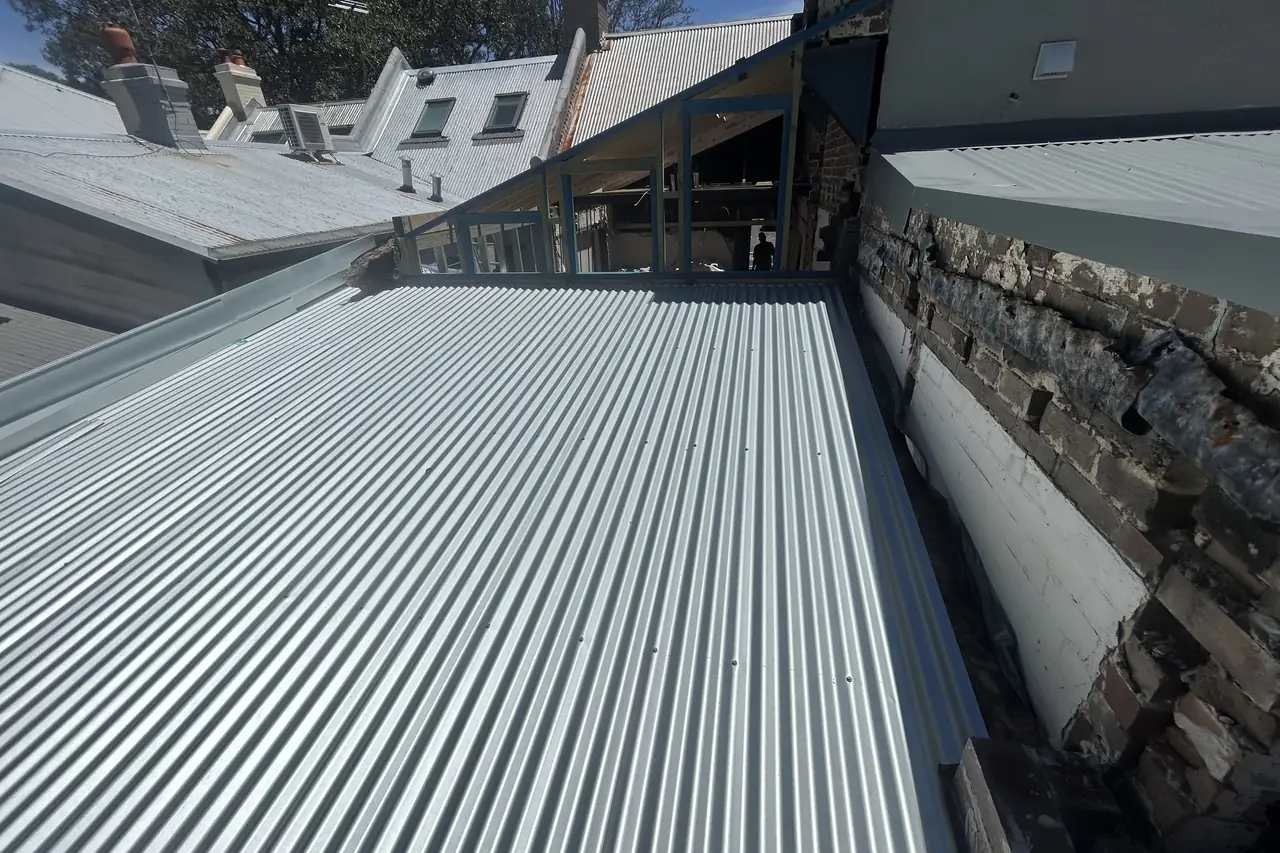 New sheeting run hard against an old rendered and stone wall, neighbouring rooftops behind
