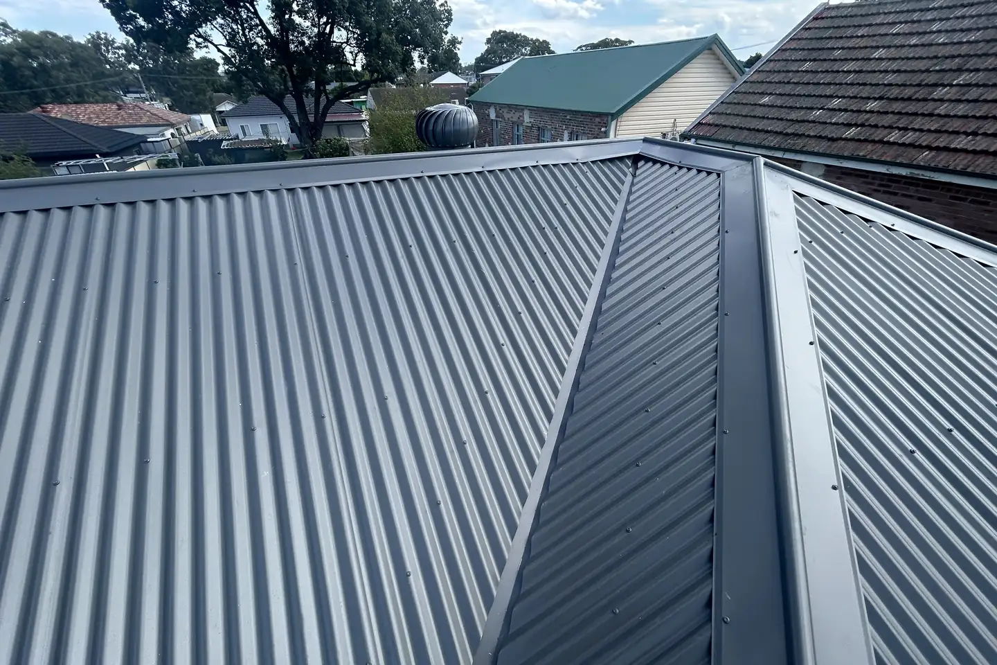 Two new steel roof planes meeting at a ridge, a whirlybird on the far slope and tiled roofs across the street