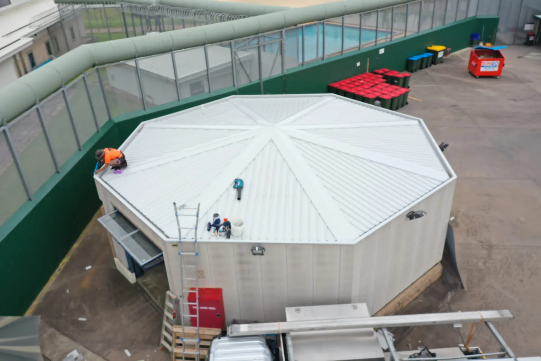 A commercial site photographed from above, with a large multi-faceted metal roof, ladders and delivery vehicles at ground level