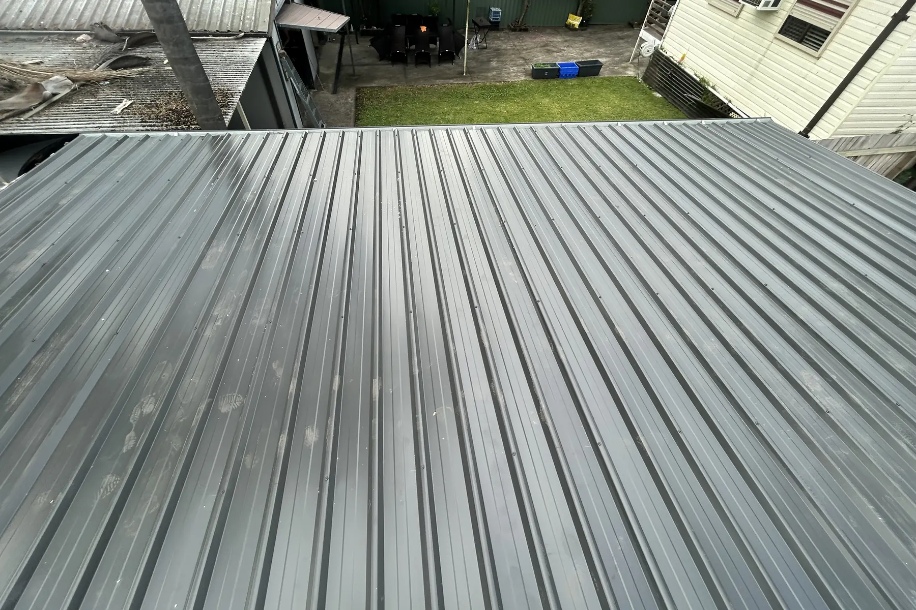 A wide low pitched roof in wide-rib steel sheeting seen from above, running out towards a backyard fence