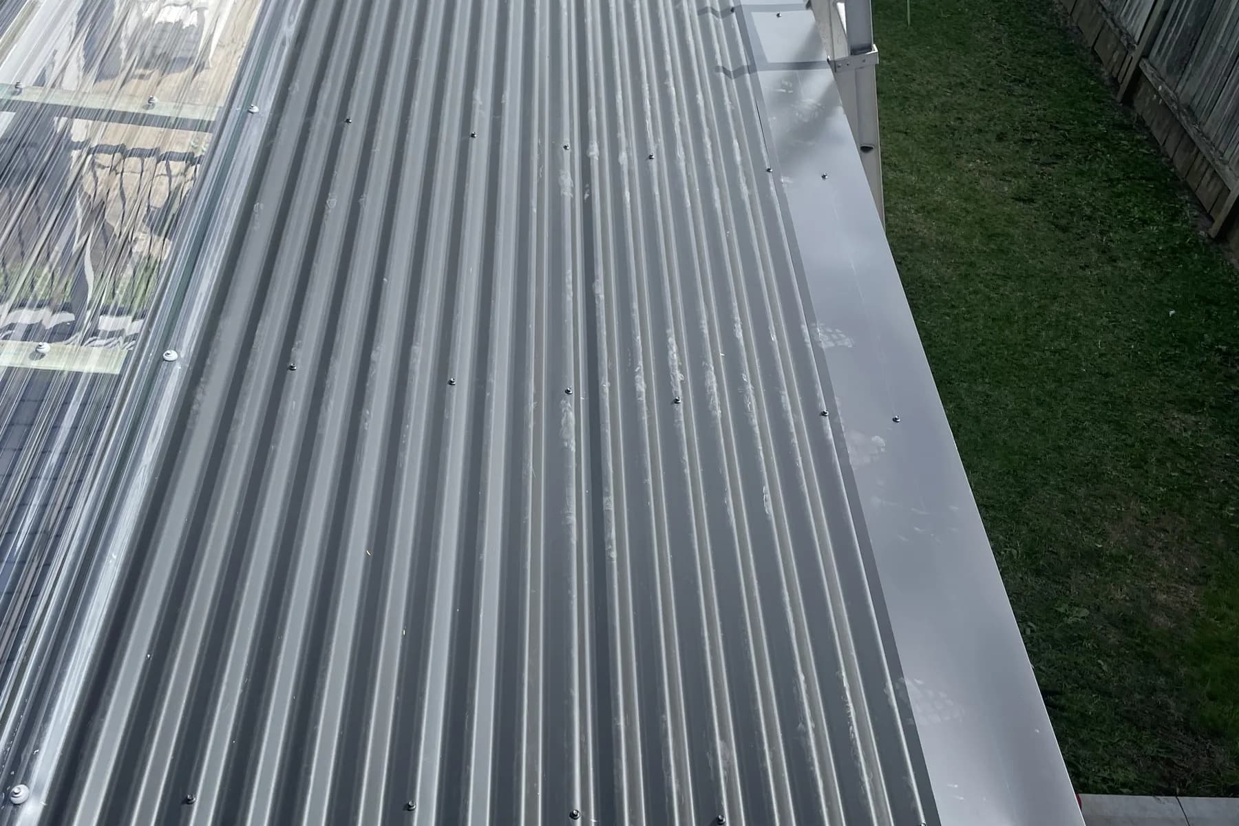 A long run of new corrugated sheeting on a low outbuilding roof, looking down over a fenced grassed yard